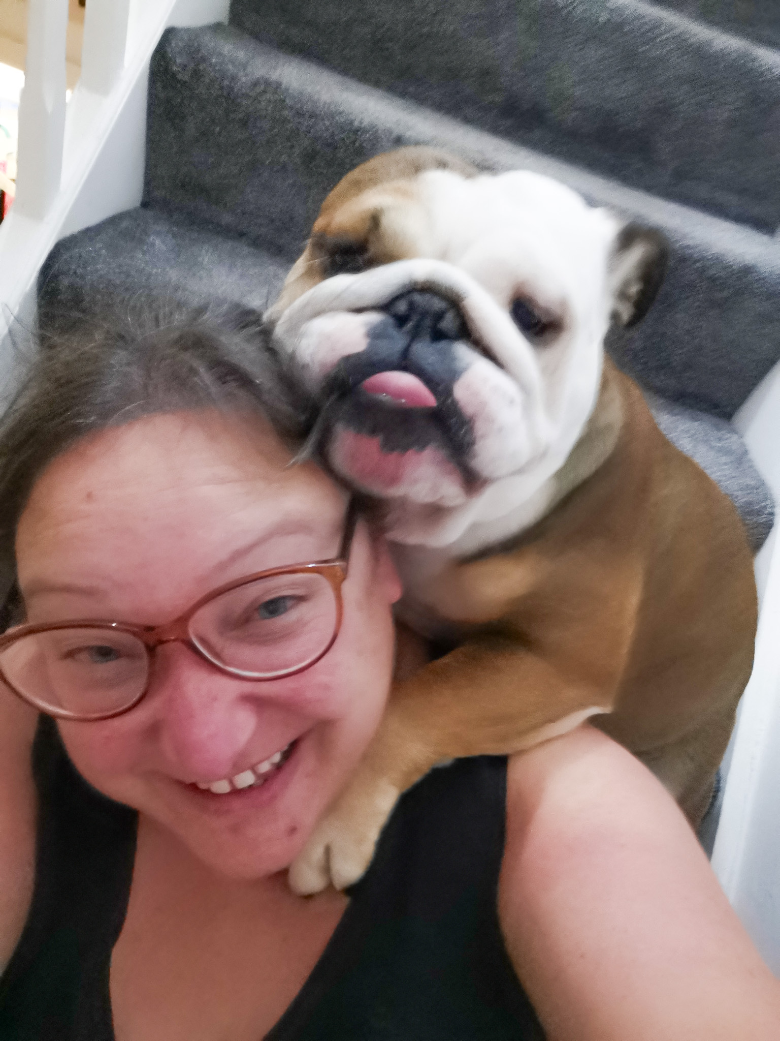 a woman wearing glasses sitting on some stairs with a tan and white coloured english bulldog on her shoulder with its tongue out.