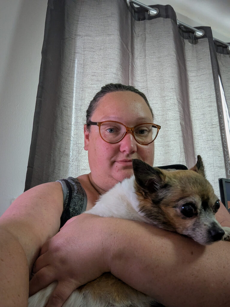 a woman wearing glasses taking a selfie with a samll tan and white coloured chihuahua in her arms who is also looking at the camera